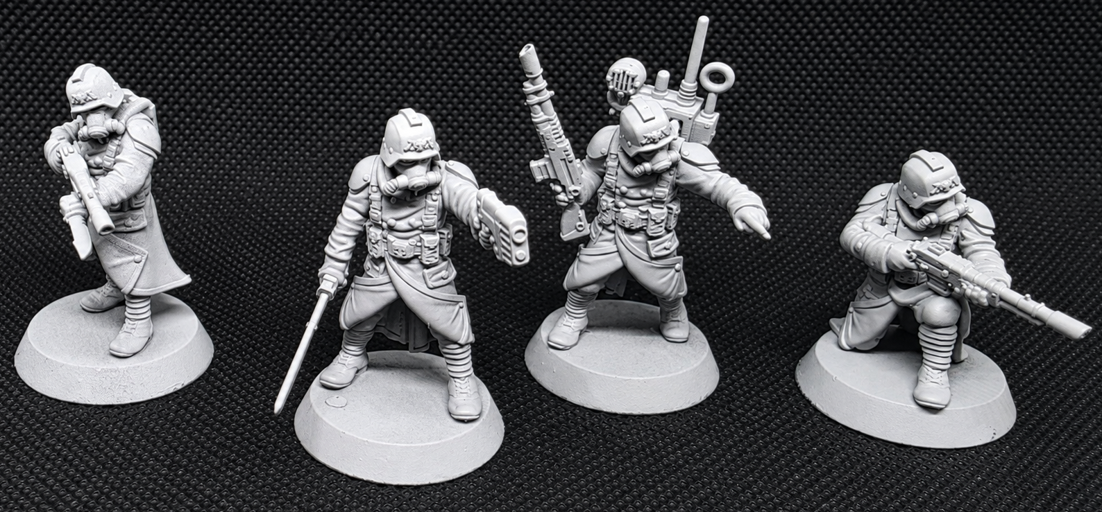 Four unpainted gray tabletop gaming miniatures in different combat poses, displayed on circular bases against a black textured background.